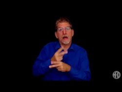 White man wearing blue shirt is standing in front of black background. He is signing "careful"