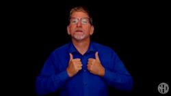 White man wearing blue shirt standing in front of black background. He is signing "residential"