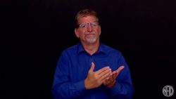 A white man with blue shirt is standing in front of a black background. He's signing "lists"