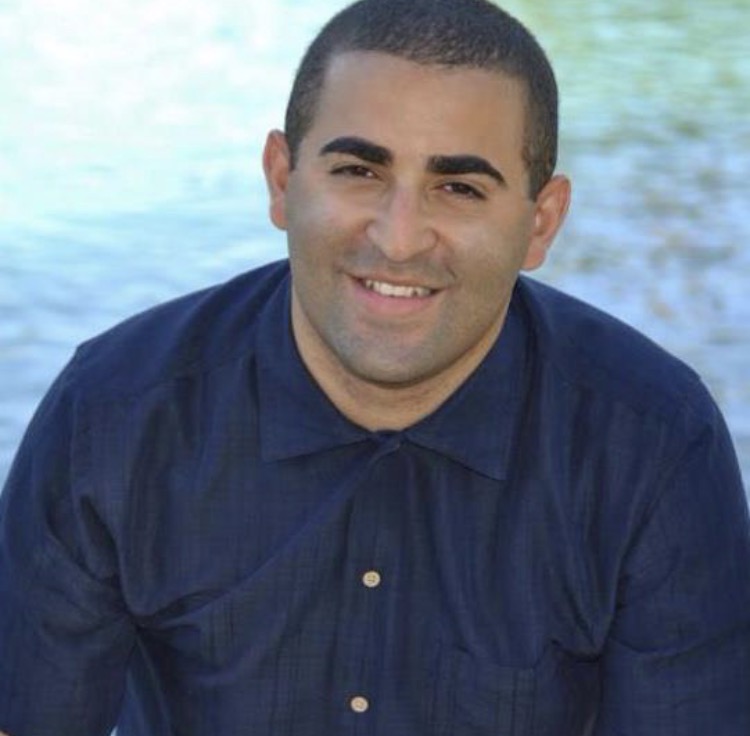 Dominican male wearing a blue buttoned down collared shirt. The image zoomed in to his head and shoulders. He sits in front of a body of water, smiles directly at the camera.
