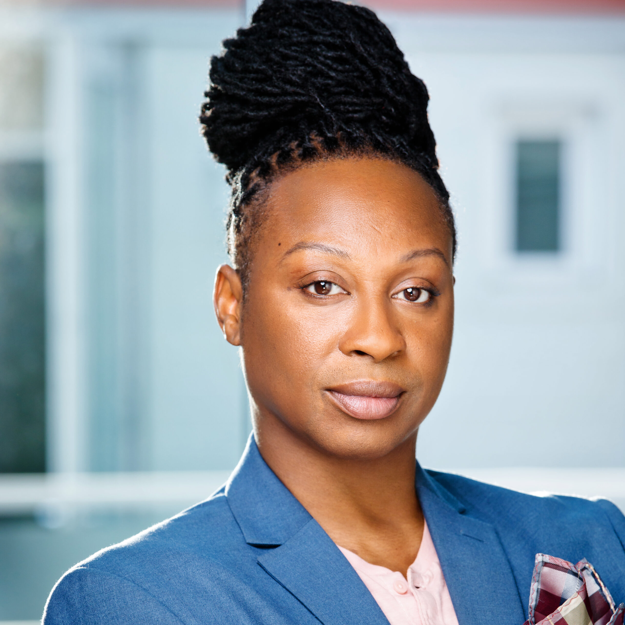 A Smoothly brown-skinned, Black Non-binary Creole African American artist, dark brown dreadlocs bun, almond dark brown eyes, wearing a baby royal suit jacket blazer, light peach shirt with three buttons.